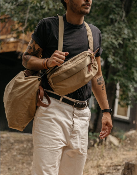Man with tattoos wears beige crossbody bag and matching tote, walking outdoors in natural light.