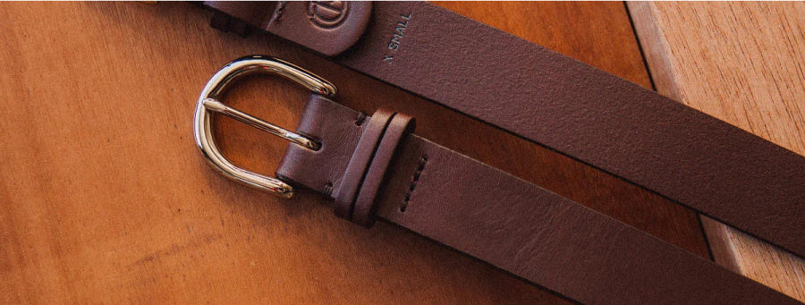 Brown leather belt with silver buckle and "X-SMALL" label, resting on wooden surface.