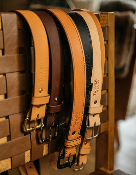 Five leather belts in tan, brown, black, and cream hang on a wooden rack, branded "TANNER GOODS," with brass and silver buckles, warm lighting.