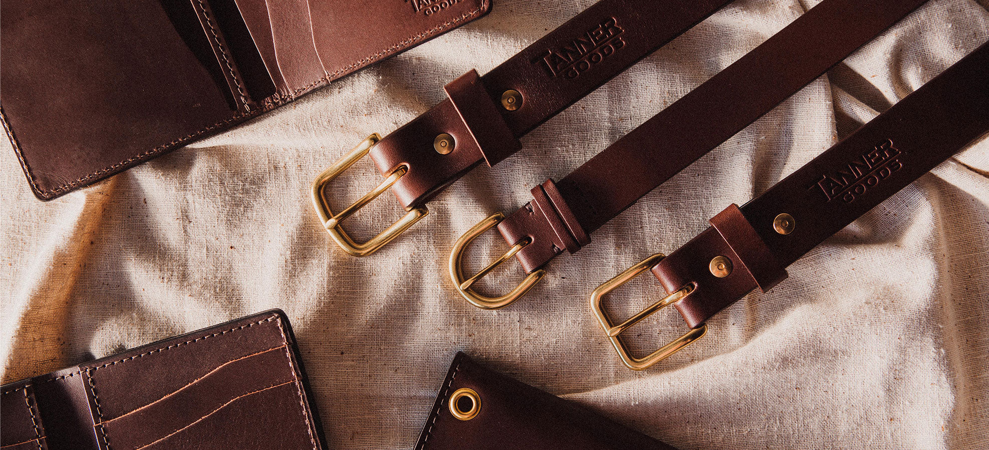 Three brown leather belts with brass buckles and "TANNER GOODS" embossed, arranged diagonally on beige fabric with matching wallets.
