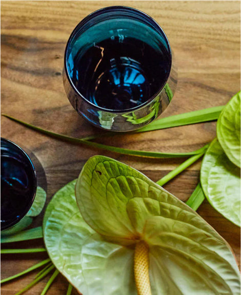 Two dark blue glass cups on wooden surface with green anthurium leaves, warm natural lighting.