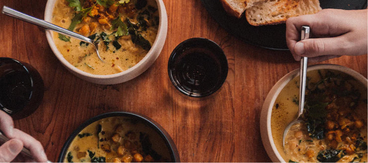 Overhead view of three bowls of creamy chickpea curry with herbs, served with bread and dark drinks on a wooden table, hands holding spoons.