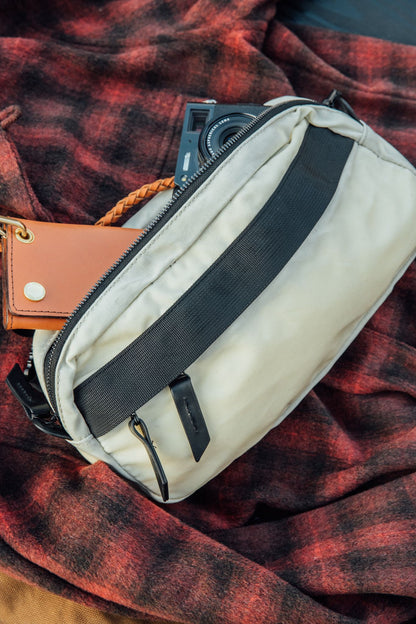 Frost waxed canvas Canyon Crossbody Pack with a black strap, partially unzipped, showing a camera and wallet, on a red plaid blanket.