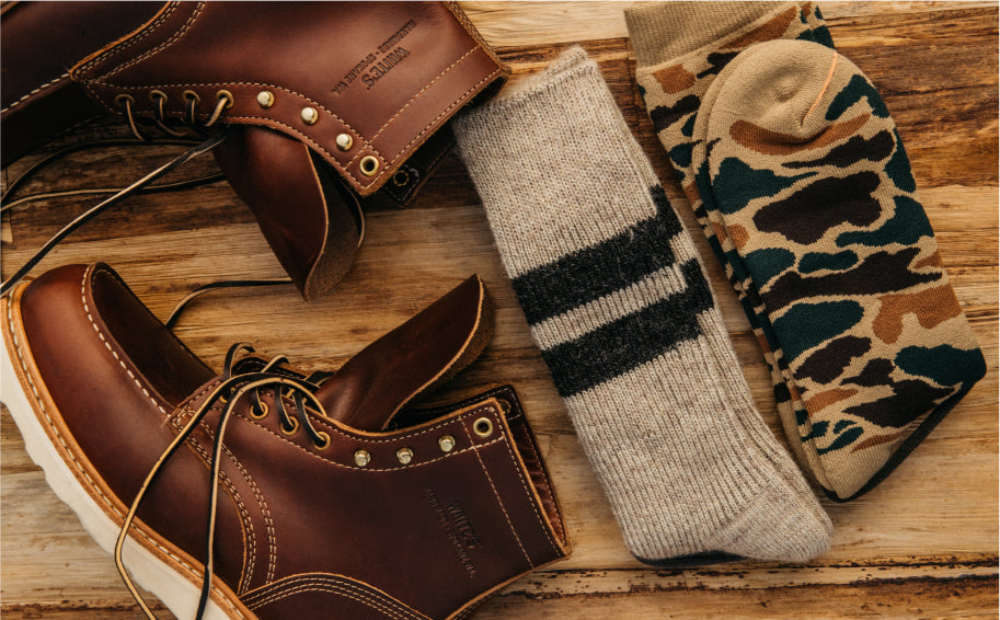 Brown leather boots with brass eyelets and white soles, paired with beige striped and camouflage socks, arranged on wooden surface.