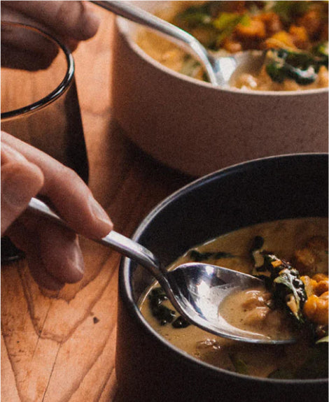 Hands holding spoons in two bowls of creamy chickpea and spinach soup on a wooden table, warm lighting.