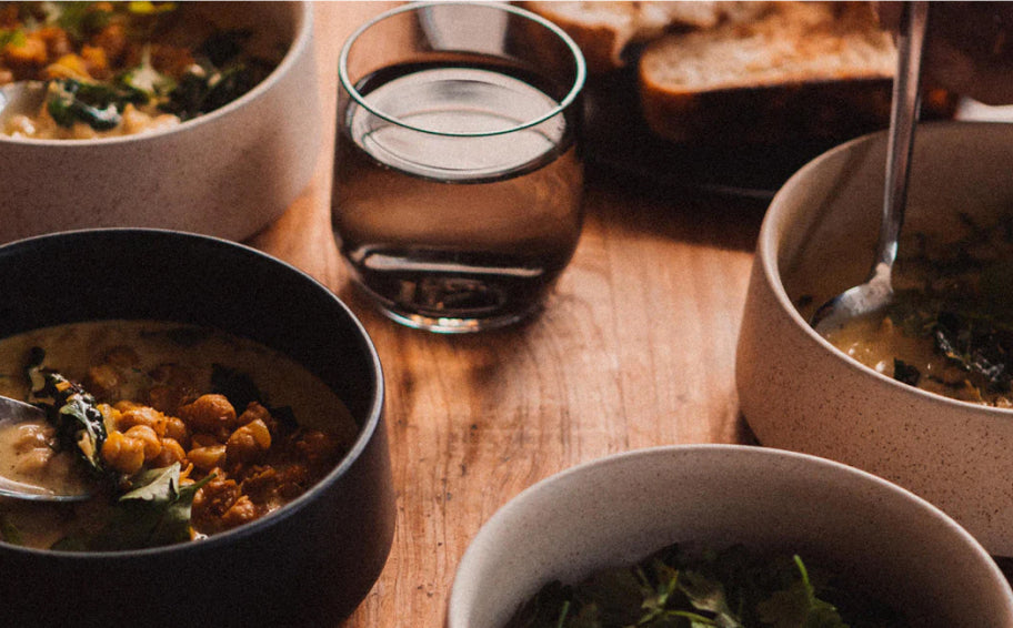 Four bowls of creamy chickpea stew with greens, a glass of water, and bread on a warm wooden table.