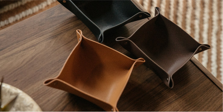Three leather desk trays in tan, brown, and black arranged on a dark wood surface with a striped rug in the background.