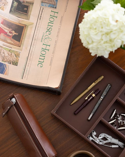 Brown leather pencil pouch with white stitching and a silver zipper, lying on a wooden desk next to a newspaper and an organizer tray.
