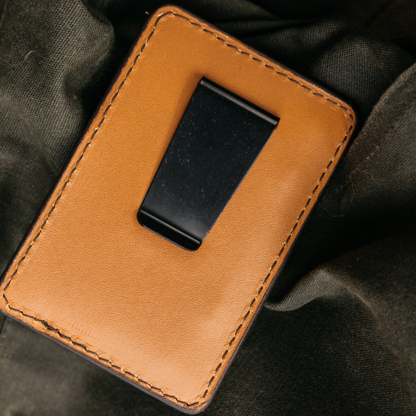 Saddle Tan leather Money Clip Wallet with a black metal clip, featuring visible stitching on a dark green fabric.