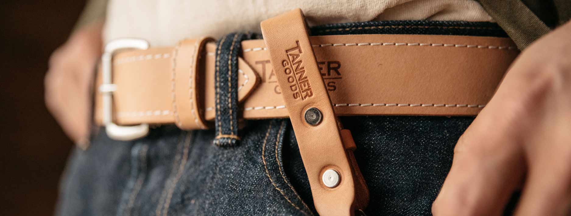 Tan leather belt with white stitching and silver buckle, branded "TANNER GOODS," worn with dark denim jeans.