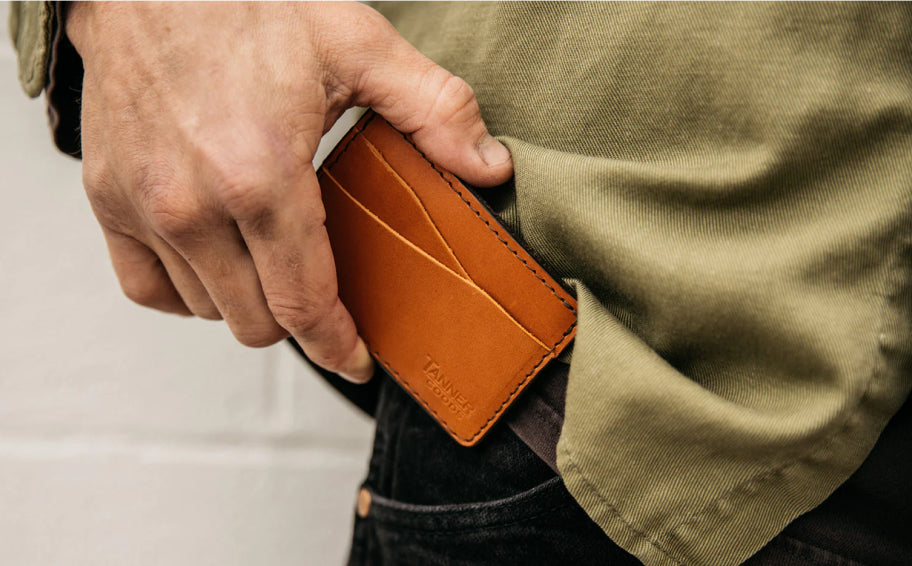 Hand placing tan leather wallet with "TANNER GOODS" embossed into front pocket of olive shirt and dark jeans.