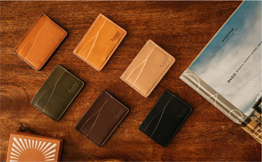 Six leather card holders in tan, brown, olive, and black on wooden surface, with book and coaster nearby.