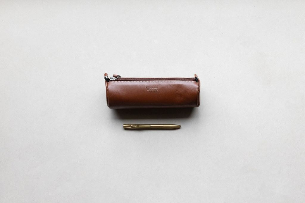 Brown leather pencil pouch with embossed text, a silver zipper, and white stitching, next to a brass pen.