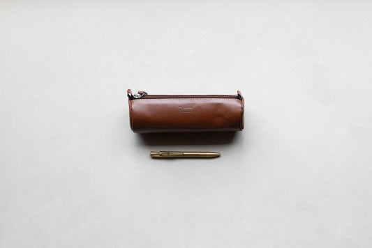 Brown leather pencil pouch with embossed text, a silver zipper, and white stitching, next to a brass pen.