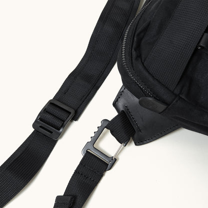 Black crossbody pack, close-up of its woven textile body, leather trim, adjustable strap with buckle and metal clip.