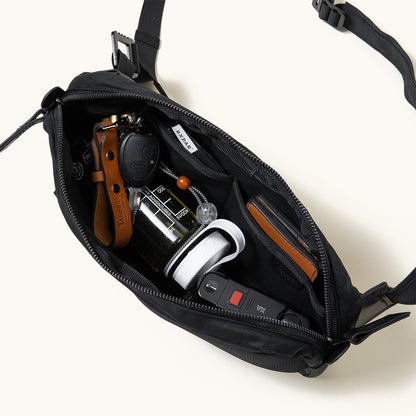 Black waxed canvas Canyon Crossbody Pack, open from above, showcasing contents like a car key, leather keychain, bottle, and wallet.