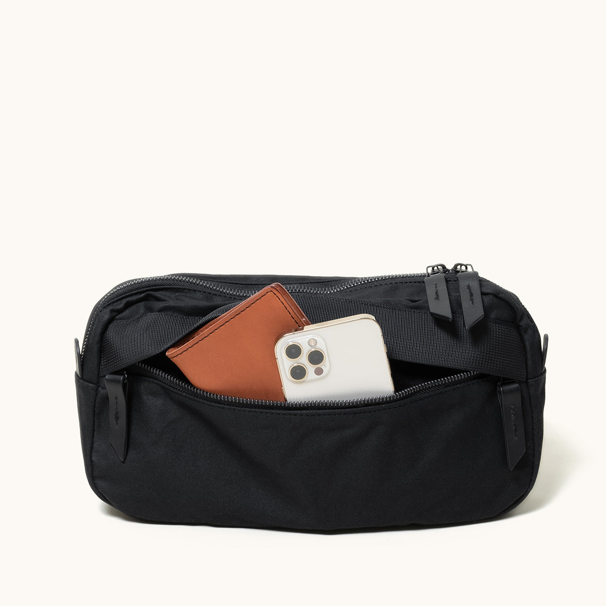 Black waxed canvas crossbody pack with an open front pocket holding a brown wallet and a light smartphone.