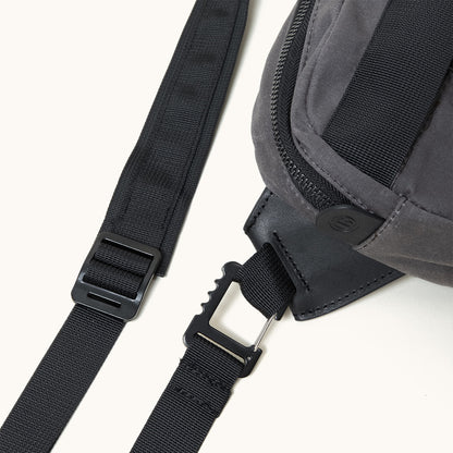 Slate grey waxed canvas crossbody pack with black nylon straps, black plastic hardware, and a debossed logo on the zipper.