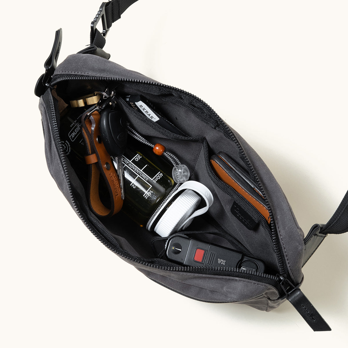 Slate grey waxed canvas crossbody pack, open from a top-down view revealing internal pockets, a water bottle, keys, and a folding knife.