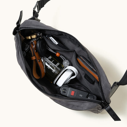 Slate grey waxed canvas crossbody pack, open from a top-down view revealing internal pockets, a water bottle, keys, and a folding knife.