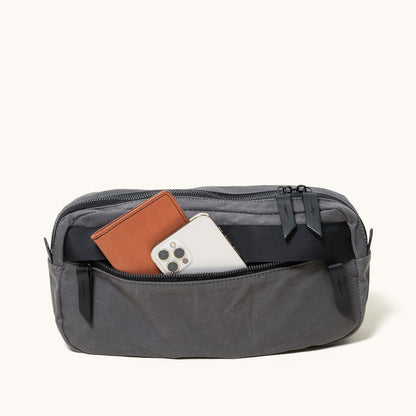 Slate Grey waxed canvas crossbody pack with an open zipper revealing a brown wallet and silver smartphone inside.