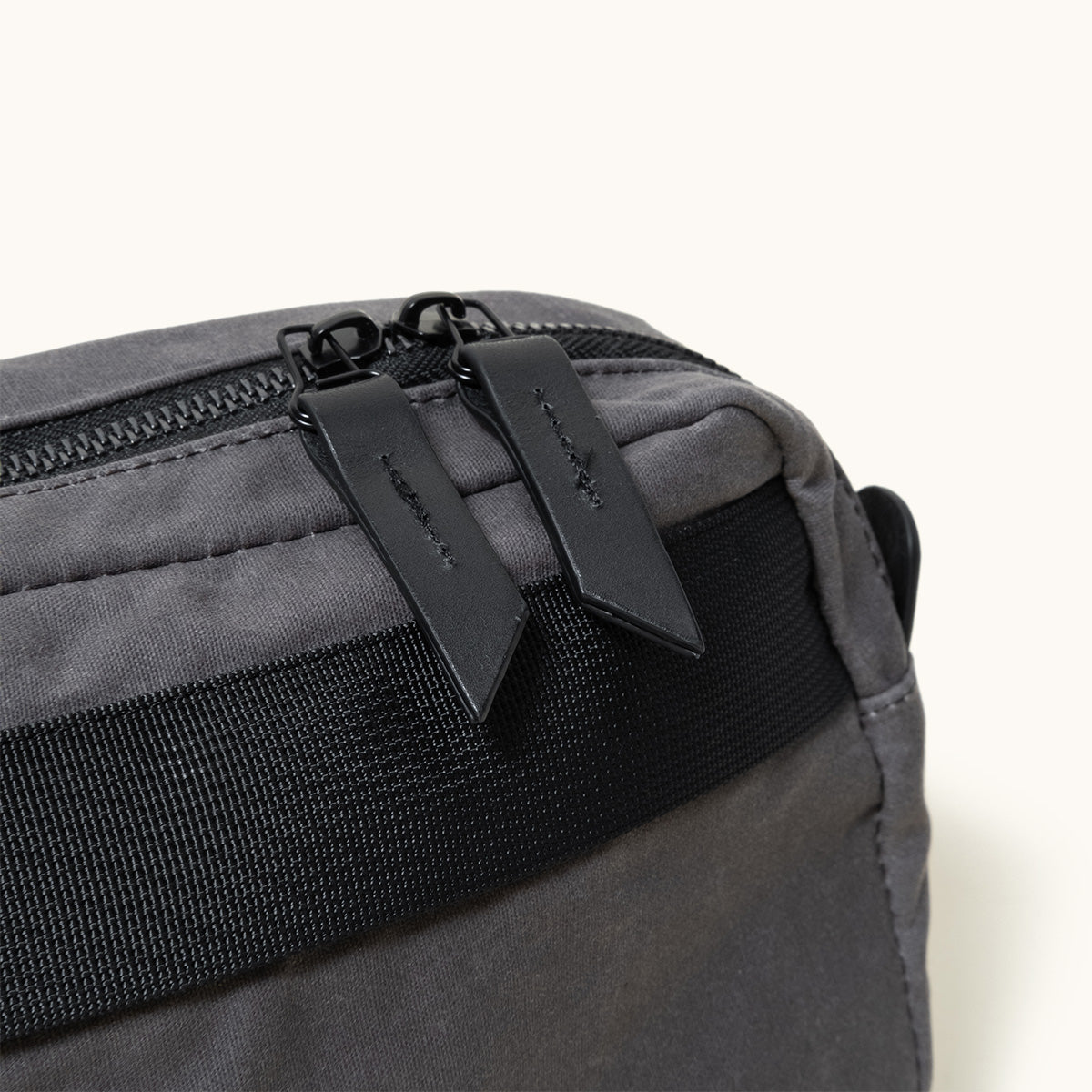 Slate grey waxed canvas crossbody pack with black zipper and two leather pulls, featuring a black woven strap.