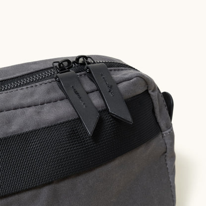 Slate grey waxed canvas crossbody pack with black zipper and two leather pulls, featuring a black woven strap.