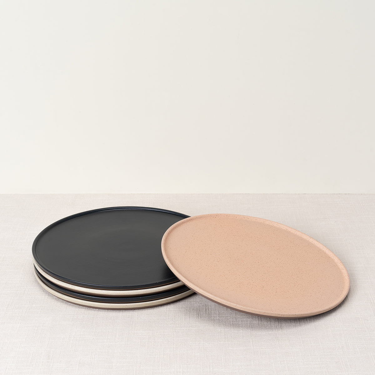 Stack of round black dinner plates and one oval speckled pink dinner plate on a light fabric surface.