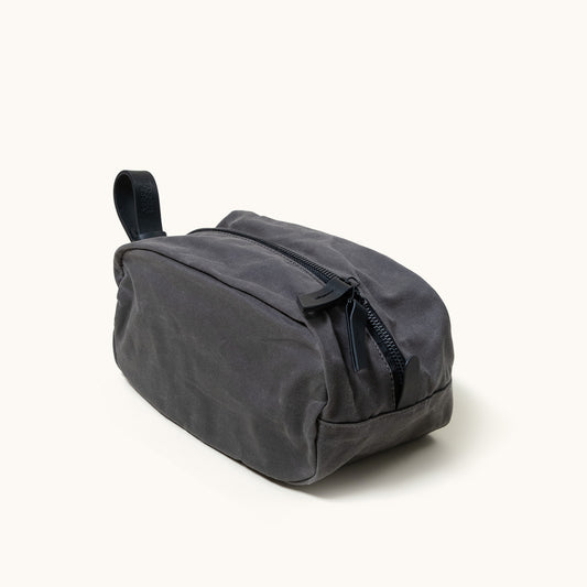 Dark grey canvas Dopp Kit with a black zipper, a black leather handle embossed with 'ZANER GOODS', angled from the side.