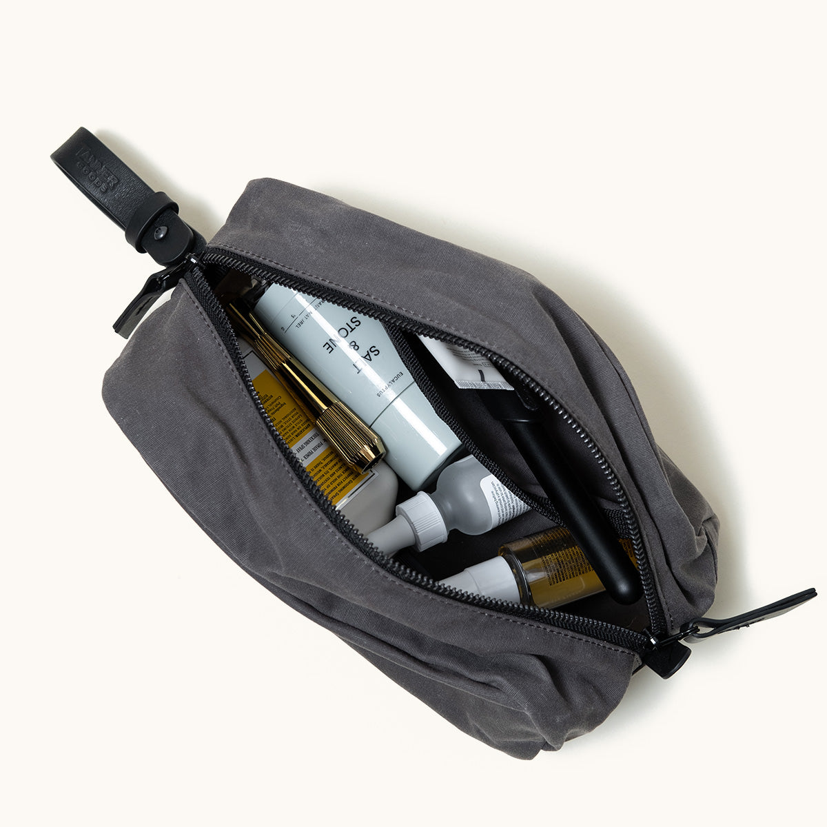 Slate grey waxed canvas Drifter Dopp Kit, open with various toiletries, black zipper, and leather strap.
