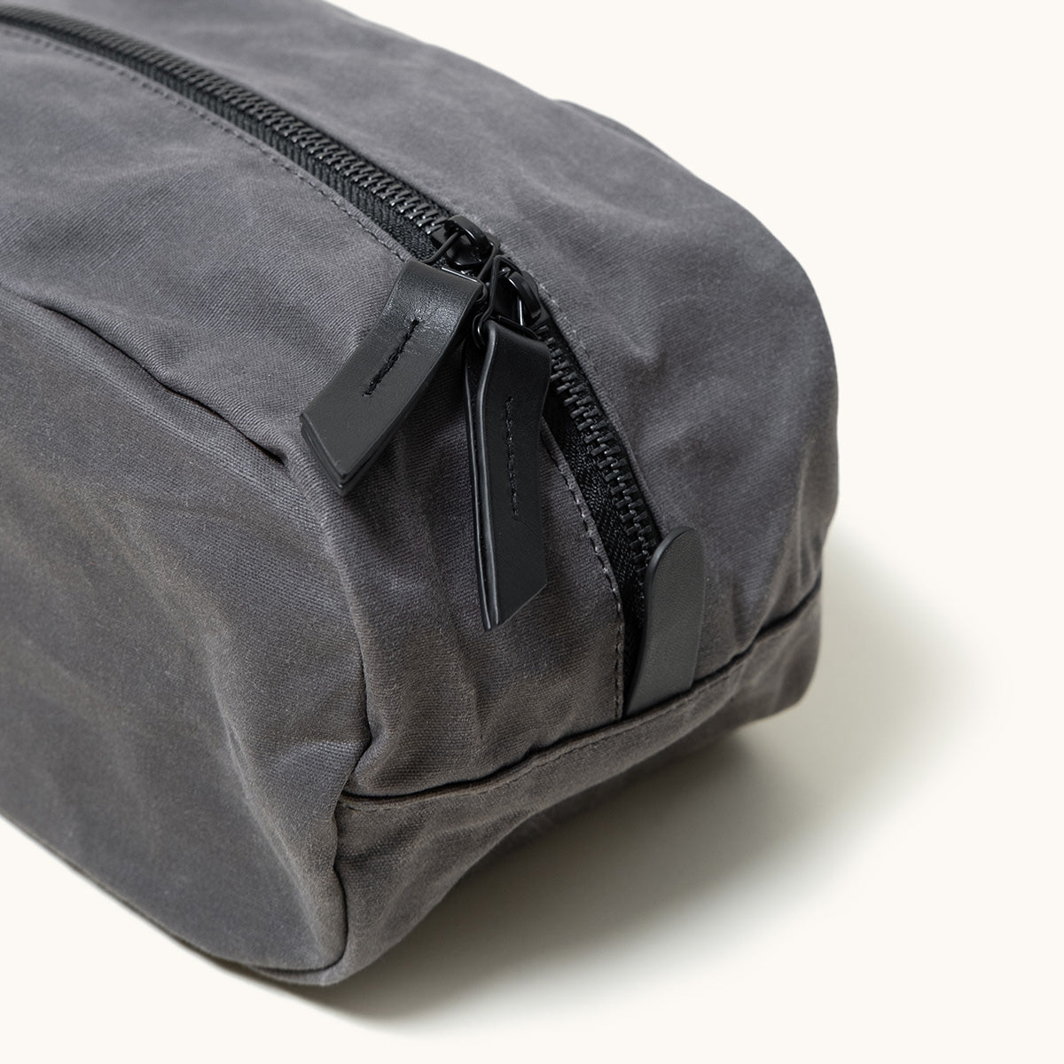 Angled close-up of a slate grey waxed canvas Drifter Dopp Kit with a black zipper and leather pulls.