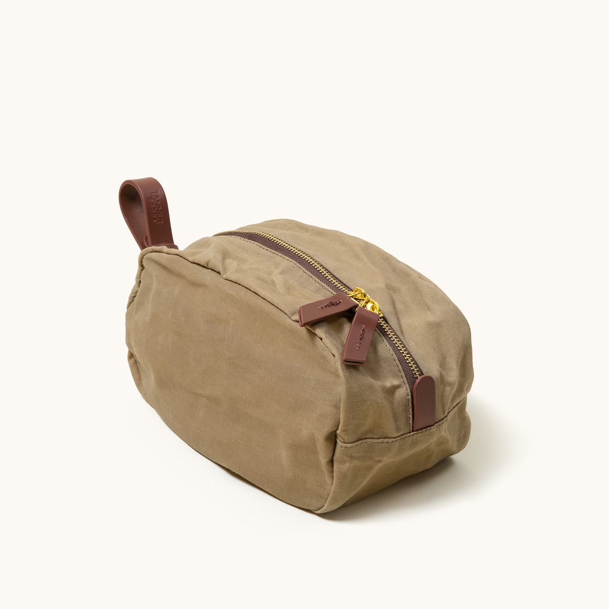 Tan canvas dopp kit with brown leather trim, a gold zipper, and a side handle, viewed at an angle.