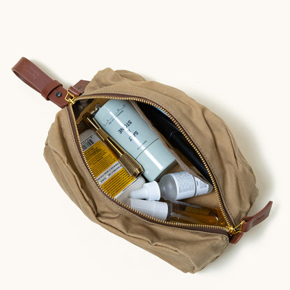 Tan waxed canvas dopp kit, open with toiletries inside, brown leather strap, and brass zipper.