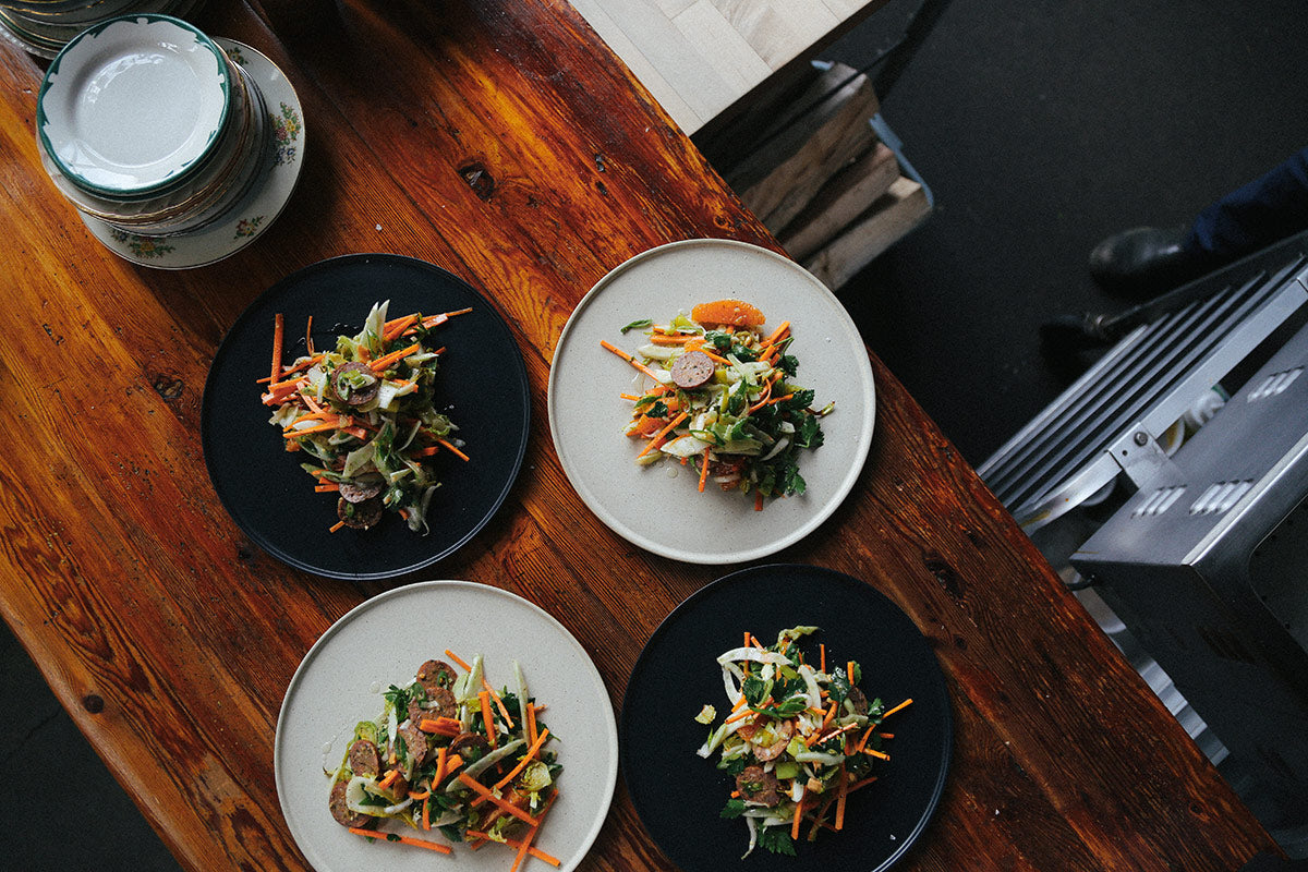 Four dinner plates, two black and two light grey, each topped with a fresh salad, on a rustic wooden table.