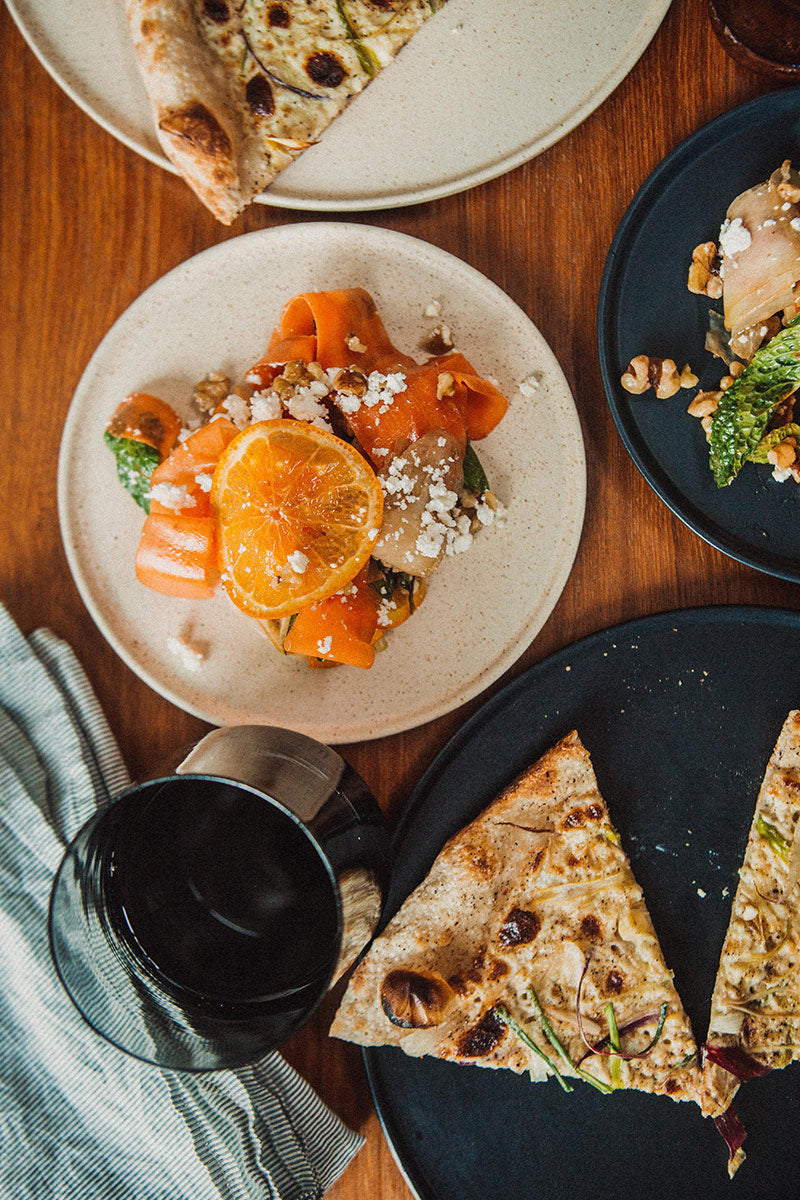 Light speckled ceramic side plates with a colorful dish of carrot ribbons and another with pizza, on a wooden table.