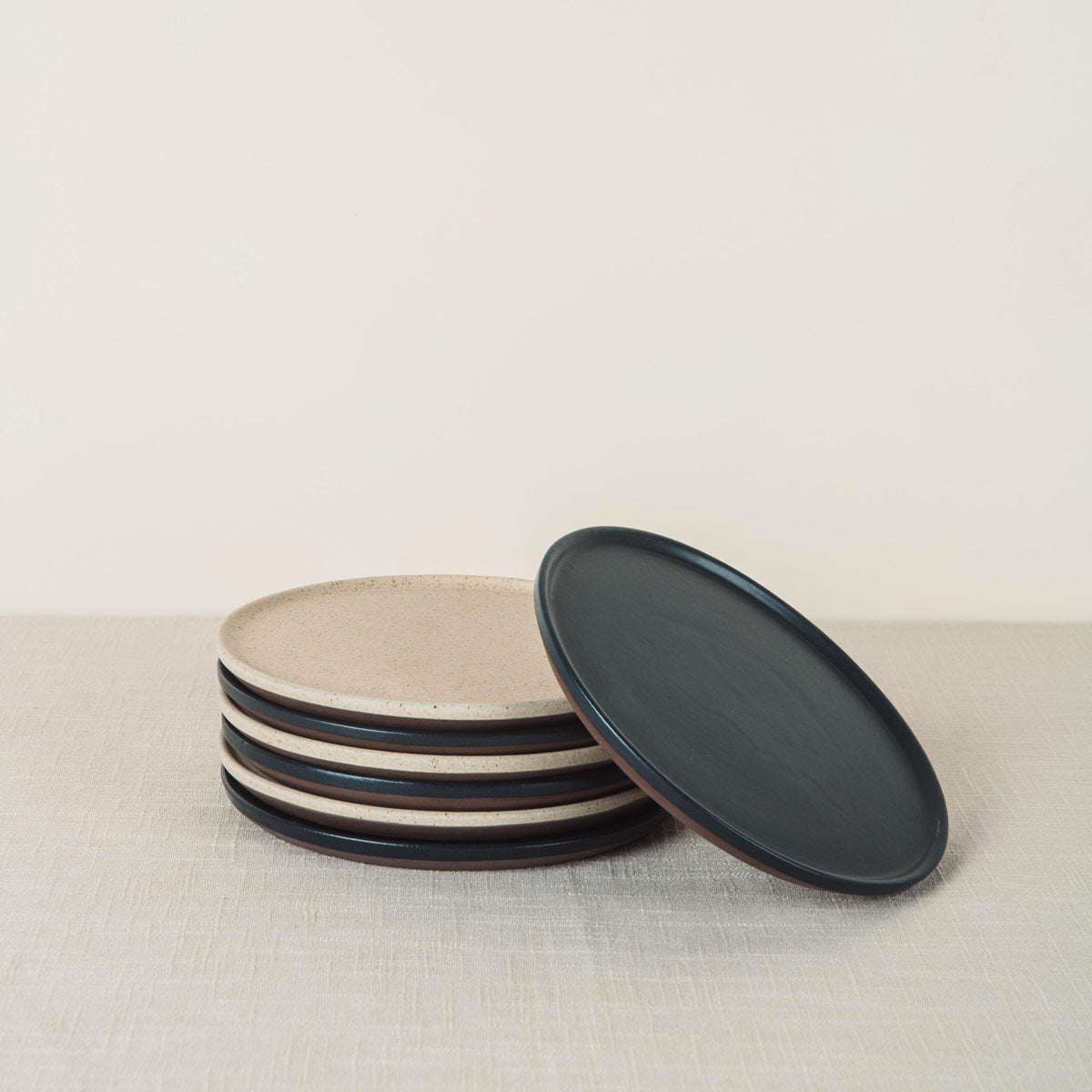 A stack of speckled beige stoneware oval side plates and one dark grey oval side plate leaning on a light tablecloth.
