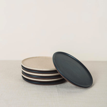 A stack of speckled beige stoneware oval side plates and one dark grey oval side plate leaning on a light tablecloth.