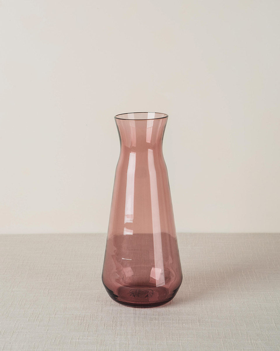 Mauve glass carafe with a tapered body, resting on a textured light beige tablecloth.