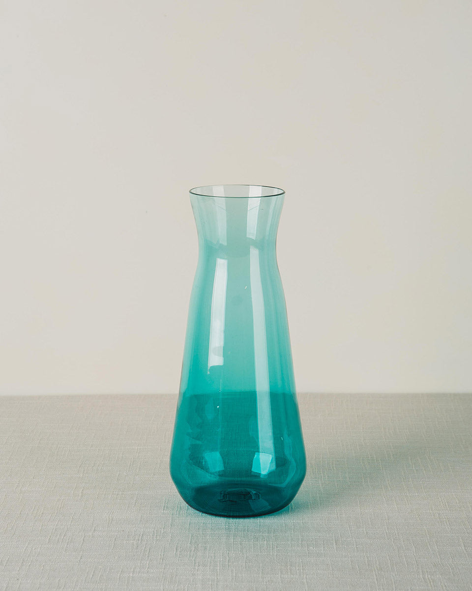 Teal glass carafe with a narrow neck and wide conical base, on a light gray textured surface.