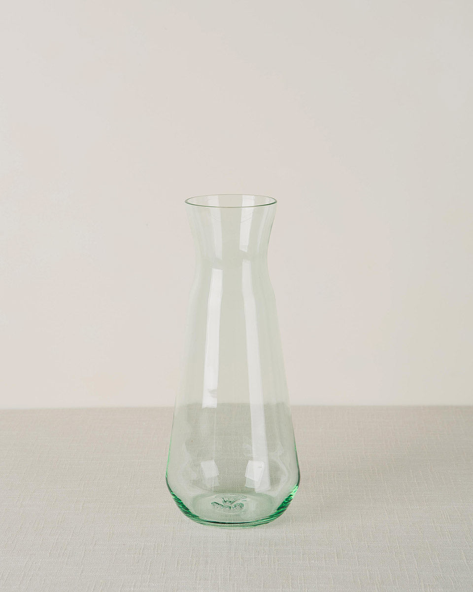 Green tinted glass carafe with a tapered body and narrow neck, on a light beige textured surface.