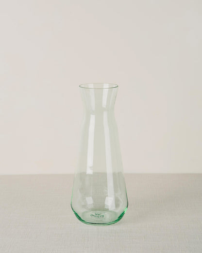 Green tinted glass carafe with a tapered body and narrow neck, on a light beige textured surface.