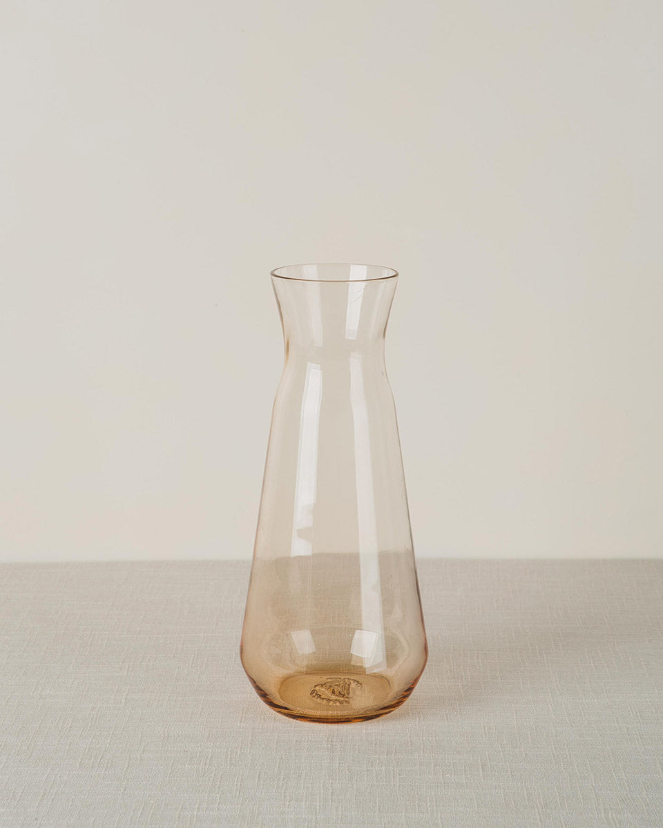 Amber-tinted glass carafe with a tapered shape and engraved detail on the base, standing on a textured light-colored tablecloth.
