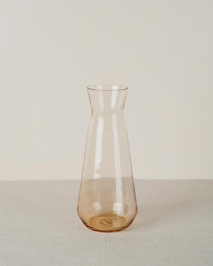 Amber-tinted glass carafe with a tapered shape and engraved detail on the base, standing on a textured light-colored tablecloth.