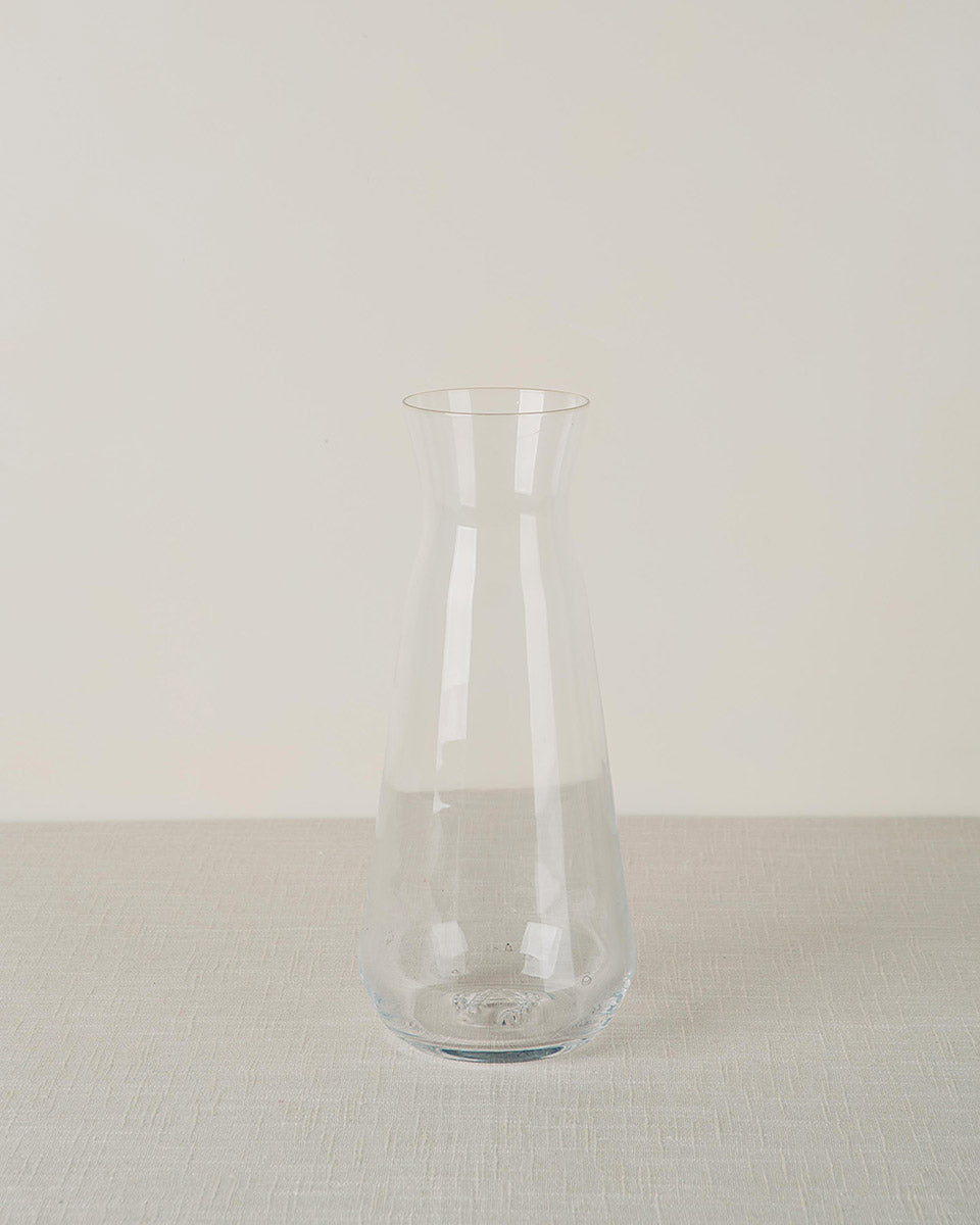 A clear glass carafe with a tapered top and bulbous base, on a light grey textured tablecloth.