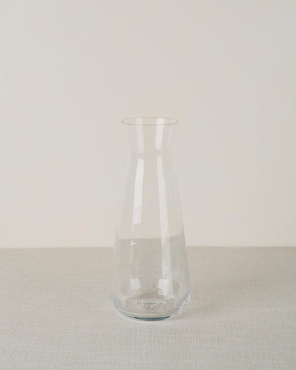 A clear glass carafe with a tapered top and bulbous base, on a light grey textured tablecloth.