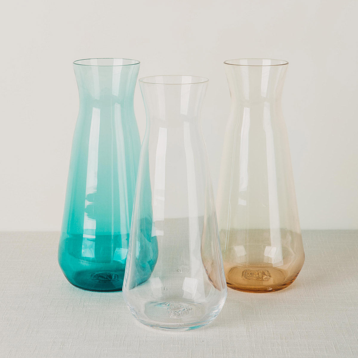 Three glass carafes, one turquoise, one clear, and one amber, standing on a light fabric surface.