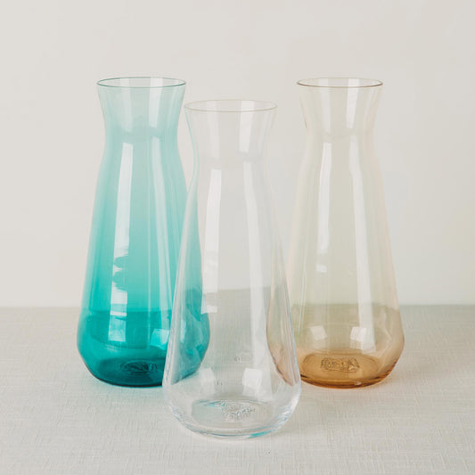 Three glass carafes, one turquoise, one clear, and one amber, standing on a light fabric surface.