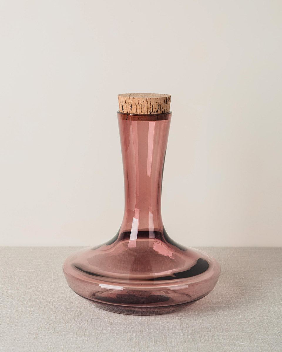 Pink tinted glass decanter with a tall neck and wide base, sealed with a cork stopper.