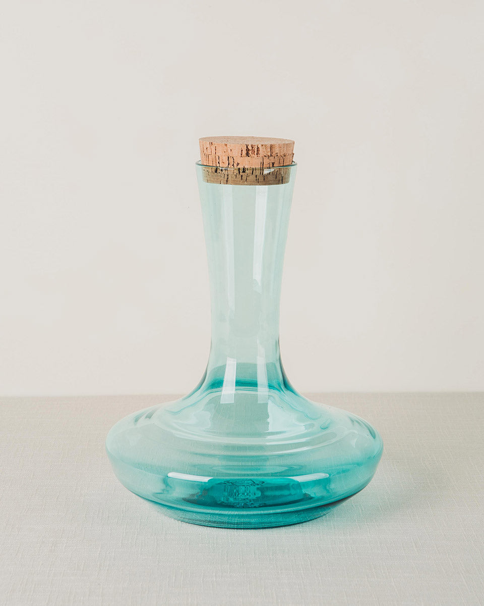 Teal glass decanter with a narrow neck, wide base, and cork stopper on a light surface.