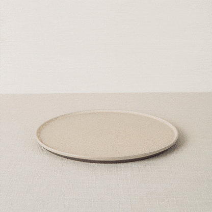 Speckled beige dinner plate with a dark brown rim, resting on a light linen surface.